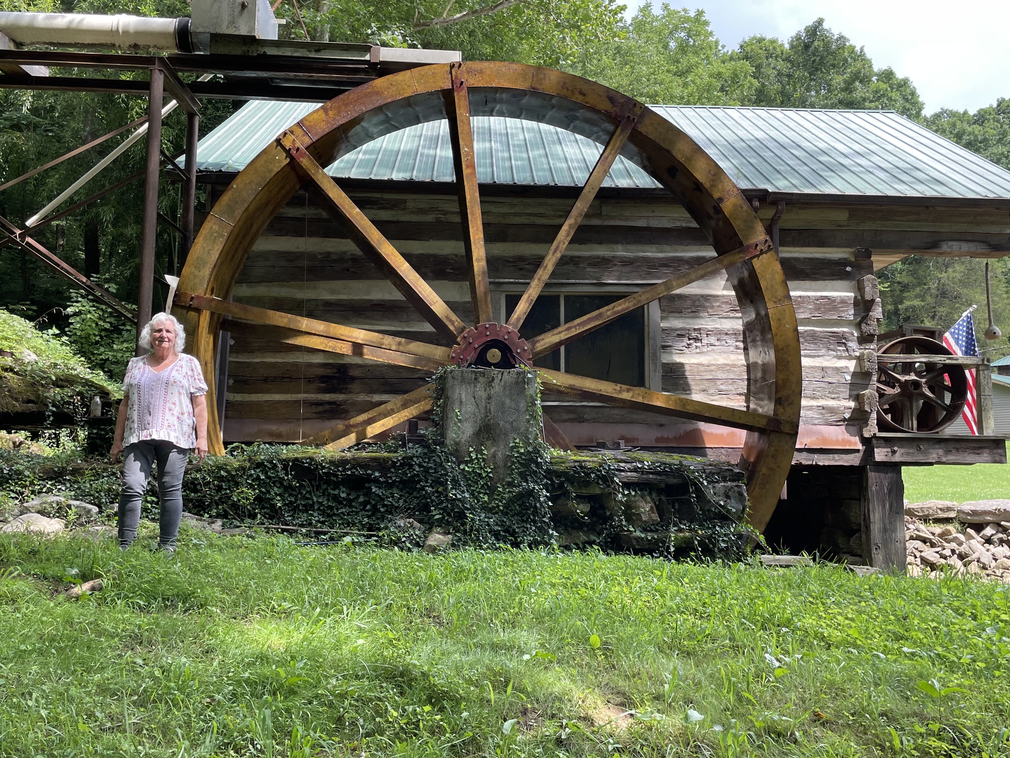 Linda standing beside a large wooden waterwheel on a day outdoors.