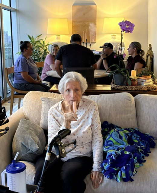 Linda seated on a sofa, enjoying a popsicle with people gathered behind her.