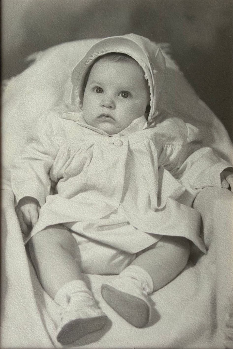 Linda as a baby wearing a bonnet and small shoes.