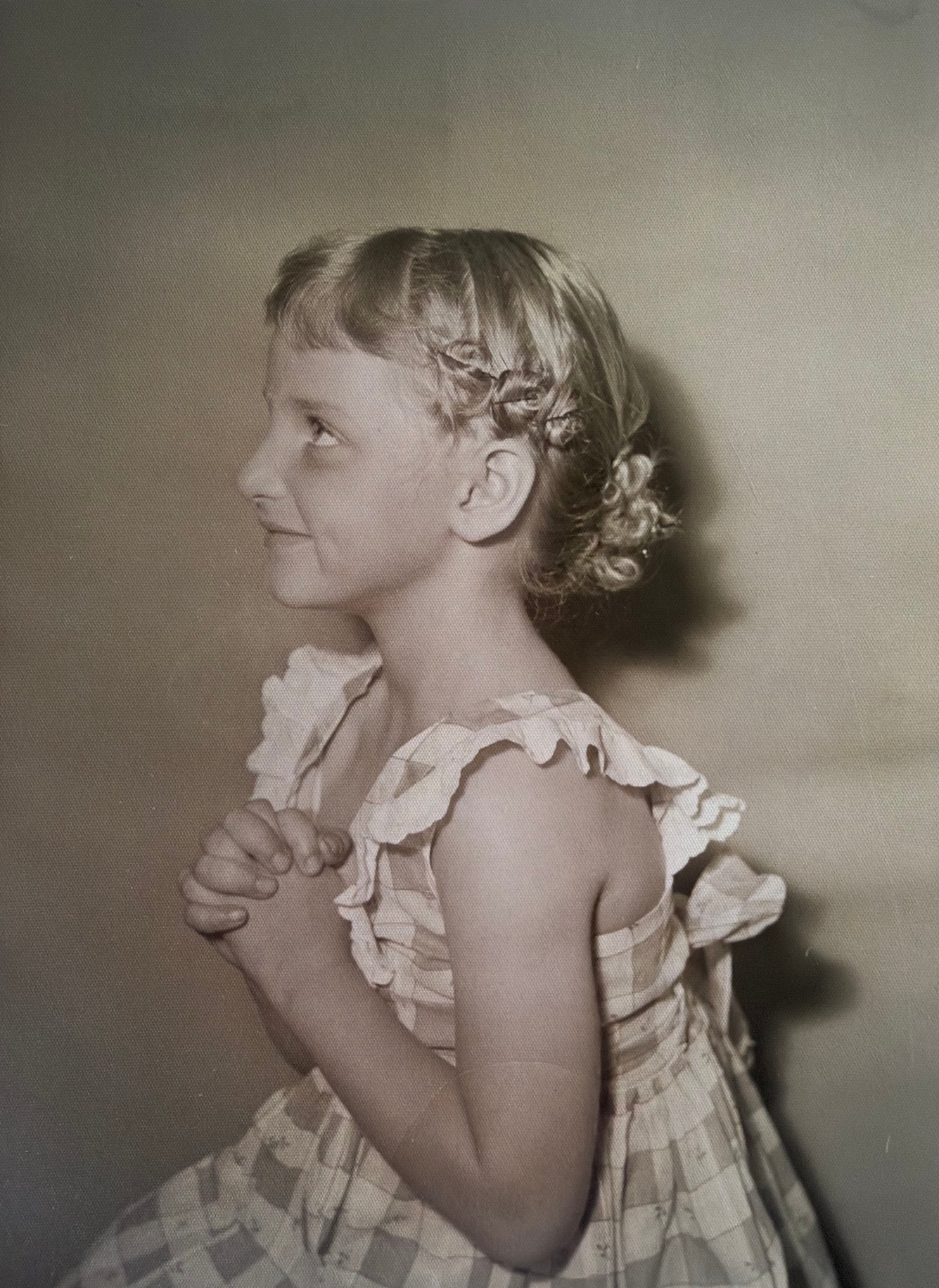 Linda as a young girl in a checked dress, looking to the side with her hands together.
