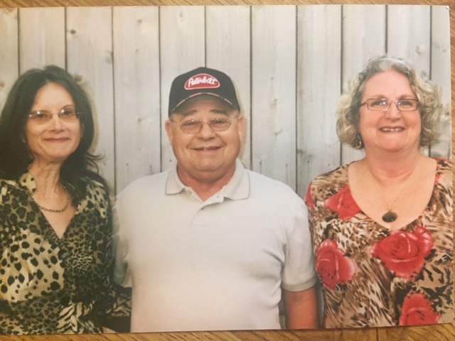 Two women and a man in a baseball cap smiling together in front of a wooden wall.